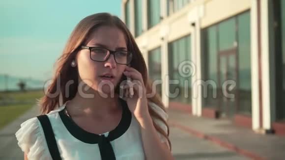 穿着白色衬衫和眼镜的女商人在一座现代化办公楼附近的街道上散步在电话里交谈视频的预览图