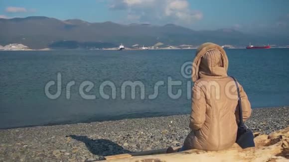 一个穿着夹克和一个包的女人坐在海边的原木上视频的预览图