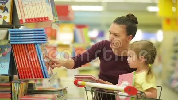 年轻的母亲和可爱的女儿在购物车选择儿童书籍在超市视频的预览图