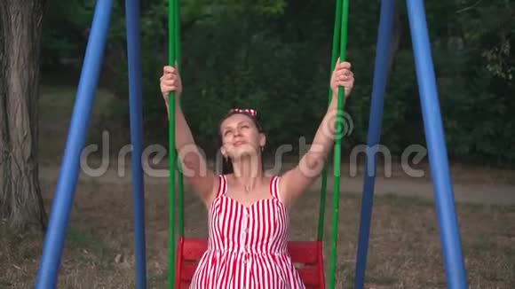 公园里的怀孕女孩在秋千上摇摆快乐的女孩一头乌黑的长发穿着条纹的白红色连衣裙在秋千上视频的预览图
