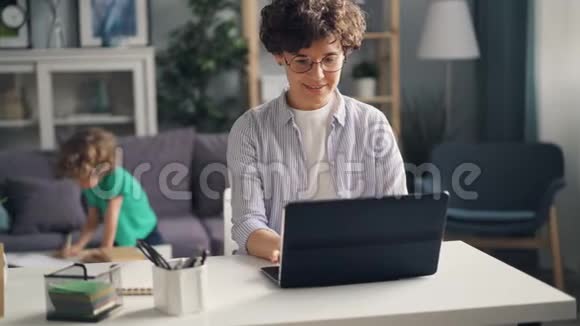 年轻女性自由职业者在家拿着笔记本电脑而小孩子在画画视频的预览图