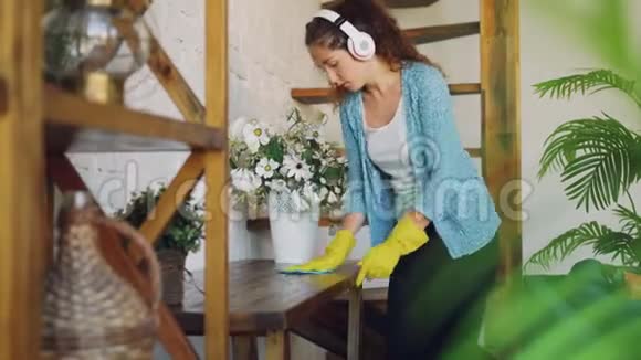 开朗的年轻女子正在家洗桌子带着耳机听音乐唱歌跳舞女孩是视频的预览图