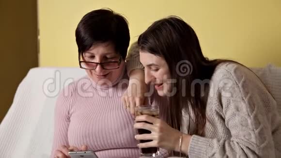 母女俩坐在家里的沙发上智能手机的使用浏览阅读新闻喝可口可乐视频的预览图