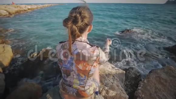 迷人的白种人女人把鹅卵石扔进大海坐在夕阳下的海岸背景上的岩石石上后方视频的预览图