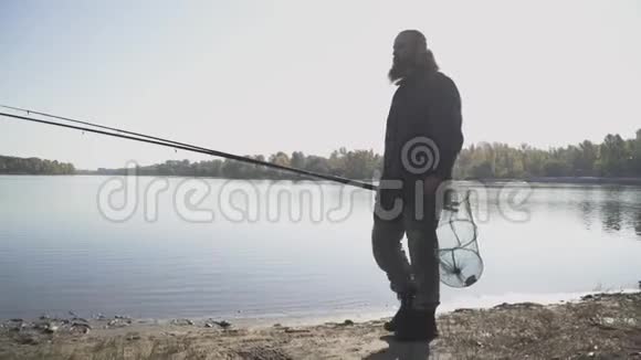 长胡子的成年渔夫带着钓竿和埋伏圈网在河边散步慢动作视频的预览图