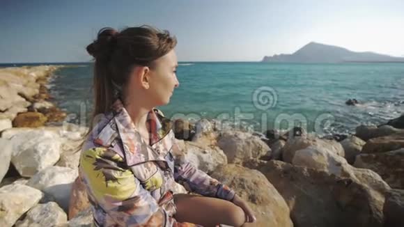 年轻迷人的女人微笑着坐在岩石上的夕阳山和海岸背景头发视频的预览图