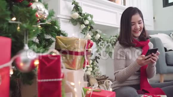 快乐的年轻亚洲女人在圣诞节期间用智能手机在家里的客厅里查看社交媒体视频的预览图