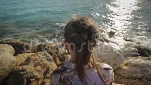 年轻迷人的女人看着夕阳坐在岩石海岸头发随风飘动后角视频的预览图