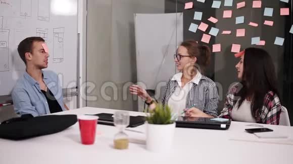 三位年轻的同事坐在现代化的阁楼办公室白色的巨大的白色桌子上一边休息一边坚持视频的预览图