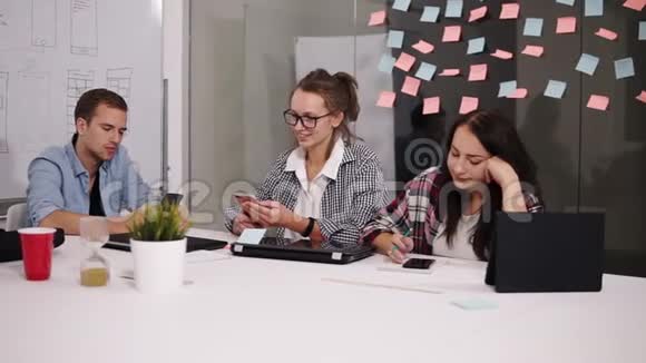 三位年轻同事坐在现代化的阁楼办公室白色的大桌子上看着他们的智能手机视频的预览图
