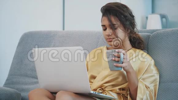 在更衣室里坐在沙发上的年轻女人他喝咖啡用笔记本电脑家庭气氛视频的预览图