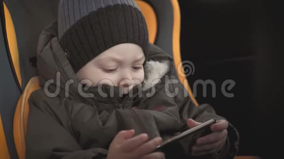 可爱的蹒跚学步的小男孩坐在汽车座椅上从智能手机上观看视频小孩在车里玩智能手机视频的预览图