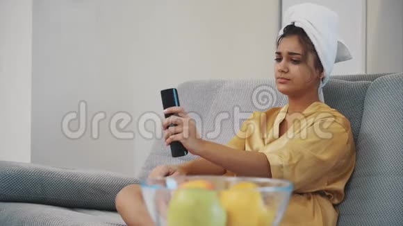 坐在沙发上的年轻女人穿着长袍头上戴着毛巾手里拿着一个黑色的控制台和一杯视频的预览图