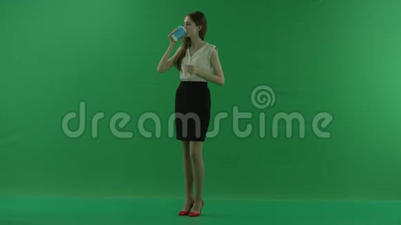 快乐的黑发年轻女子正拿着一杯咖啡或茶喝着她侧身站在绿色的屏幕上视频的预览图