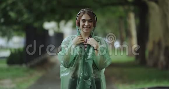 年轻的女士站在雨中微笑着听耳机里的音乐穿着雨衣视频的预览图