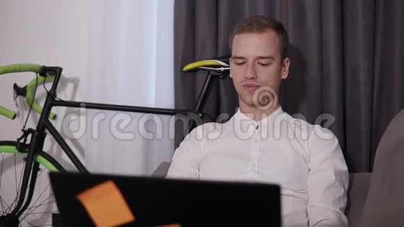 一个年轻的金发男子穿着白色衬衫坐在家里的沙发上工作或玩耍的正面景色刺激视频的预览图