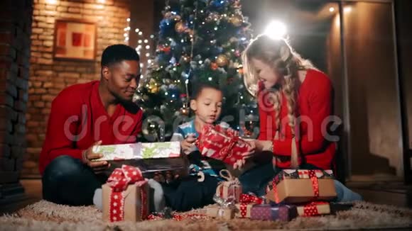 新年礼物中一个幸福的家庭正坐在圣诞树附近那个男孩正在用红色包装摇动礼物视频的预览图