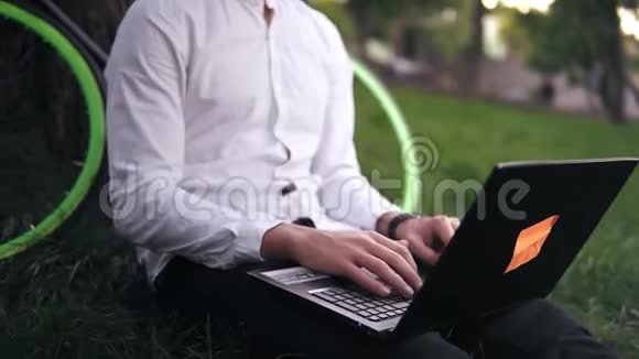 年轻的成功商人穿着白色衬衫和黑色眼镜微笑的人坐在草地上在城市的笔记本电脑上工作视频的预览图