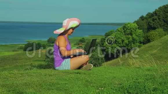 一个戴着草帽的女人坐在山上俯瞰湖面在笔记本电脑上排版文字视频的预览图