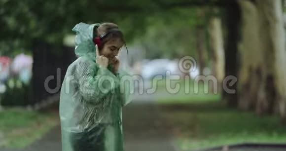 漂亮的年轻女人在下雨天在公园里跳舞听着耳机里的音乐穿着雨衣视频的预览图