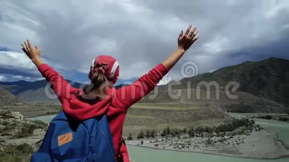 年轻快乐的旅行女孩带着背包和帽子站在山的边缘把他的手放在空中回去视频的预览图