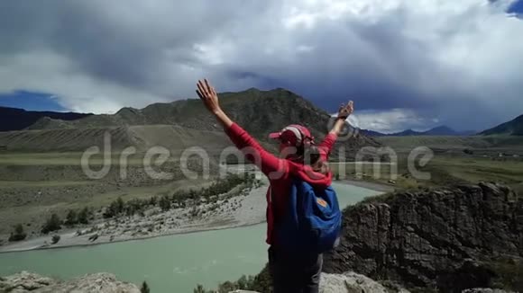 年轻快乐的托勒斯特女人带着背包和帽子站在山的边缘把他的手举在空中回去视频的预览图