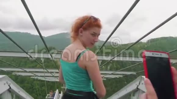 两个年轻的女人在青山谷和吊桥的背景上互相拍照高视野视频的预览图