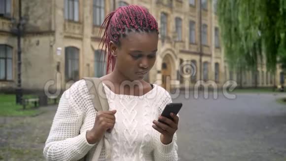年轻的非洲女人带着粉红色的可怕的袜子打电话拿着背包站在大学附近的公园里快乐视频的预览图
