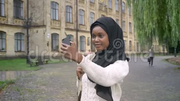 年轻的非洲穆斯林妇女戴着头巾带着手机自拍站在大学附近的公园里迷人的女性视频的预览图
