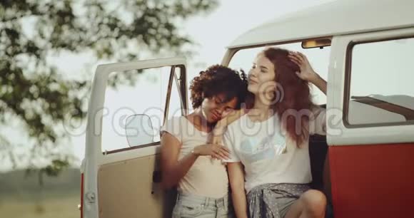 两位卷发的年轻女士坐在老式橙色巴士旁边微笑着看着对方视频的预览图