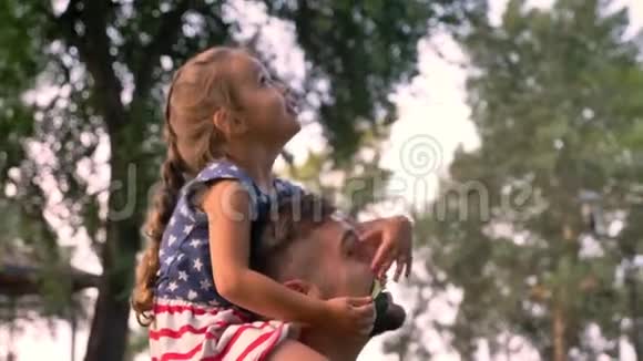 年轻的一家人夏天在公园散步女孩坐在父亲肩膀上他们看着树视频的预览图
