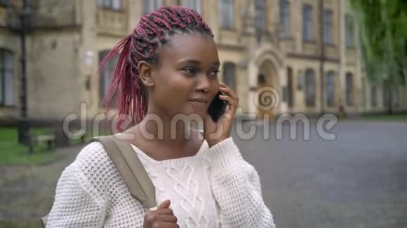 年轻的非洲漂亮学生在电话里谈笑风生背着背包站在大学附近的公园里视频的预览图