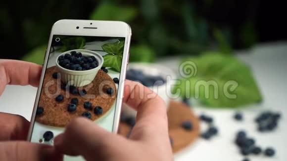 女孩在白色桌子上的白色碗中拍摄美味蓝莓的照片背景是绿色植物视频的预览图