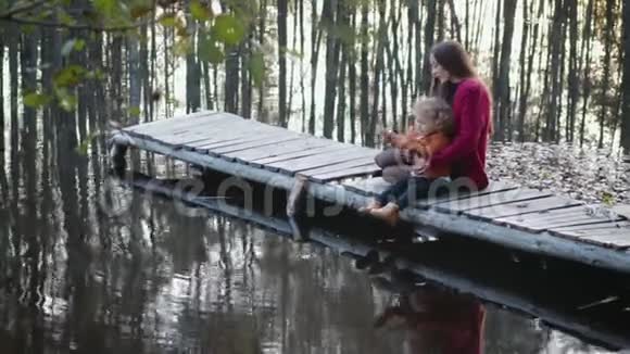 母子俩坐在湖边的一座木桥上欣赏美丽的景色家庭的概念视频的预览图