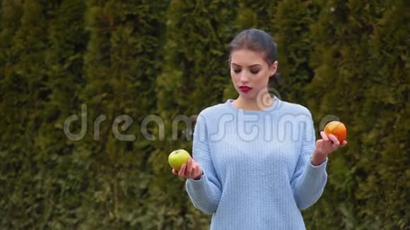 在绿苹果和甜橙之间穿着蓝色夹克的漂亮年轻女子选择了肖像决定咬苹果视频的预览图