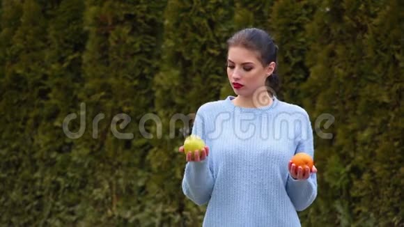 在绿苹果和甜橙之间穿着蓝色夹克的漂亮年轻女子选择了肖像决定咬苹果视频的预览图
