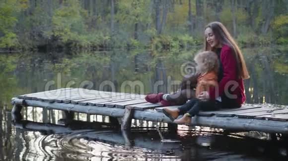 母子俩坐在湖边的一座木桥上欣赏美丽的景色家庭的概念视频的预览图