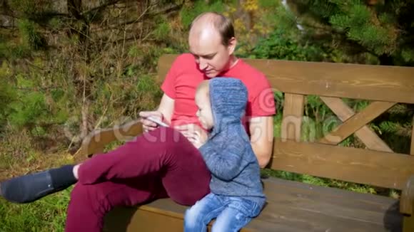 一个有孩子的男人坐在长凳上用智能手机父子俩笑眯眯的看着屏幕上的小玩意4k视频的预览图