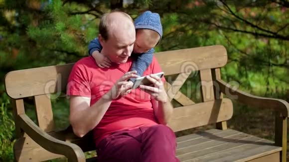 这个男孩用智能手机拥抱他的父亲一个男人带着孩子坐在长凳上的智能手机上拍照视频的预览图