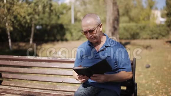 一位戴眼镜的老人在公园长椅上看电子书视频的预览图