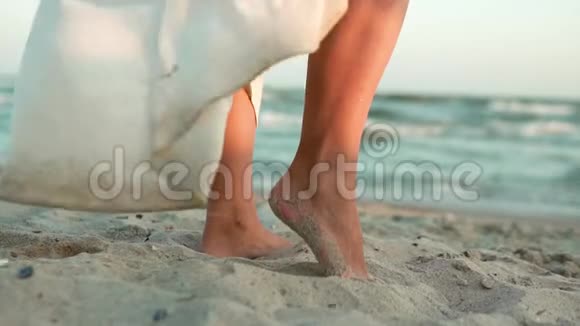 一个穿着白色长裙的白种人女孩的腿站在蔚蓝的大海的岸边站在沙滩上太阳镜头慢慢视频的预览图