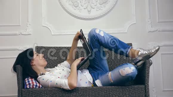 年轻的女人女孩黑发穿着白色衬衫和牛仔裤躺在沙发上小沙发带笔记本电脑听音乐视频的预览图