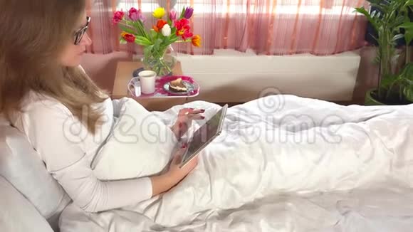 女人坐在床上用平板电脑早餐和鲜花放在橱柜里视频的预览图