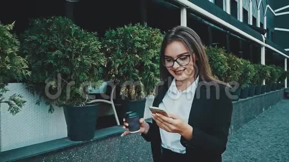 年轻漂亮聪明的商务女孩穿着商务服走路在这段时间里她喝咖啡看着电话视频的预览图