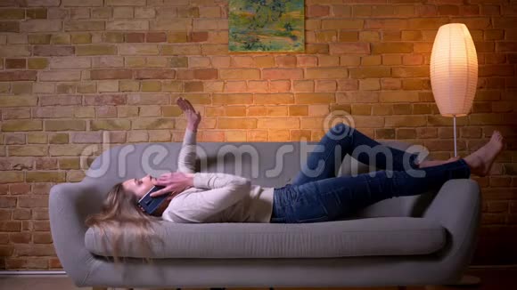 一位年轻迷人的白种人深色头发的女性躺在室内的沙发上打着一个电话视频的预览图