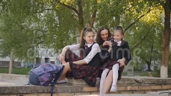 母亲带着两个穿校服的女儿背包坐在学校附近的街道上亲吻和交谈视频的预览图