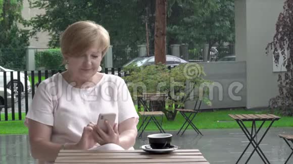 年老的女人坐在露台上滚动电话下大雨视频的预览图