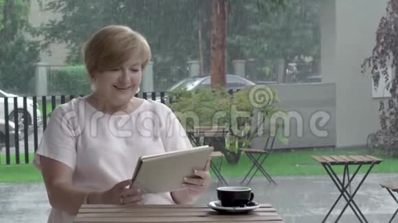 快乐的老妇人一边微笑着看着平板电脑一边思考一边打字坐在夏天的露台上视频的预览图