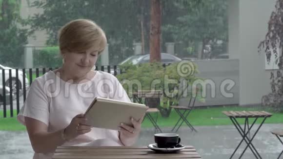 一位有魅力的老妇人拿着一杯茶在露台上的平板电脑上看书和打字视频的预览图
