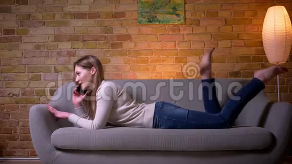 一位年轻迷人的白种人深色头发的女性在一个舒适的室内沙发上躺着一个电话视频的预览图
